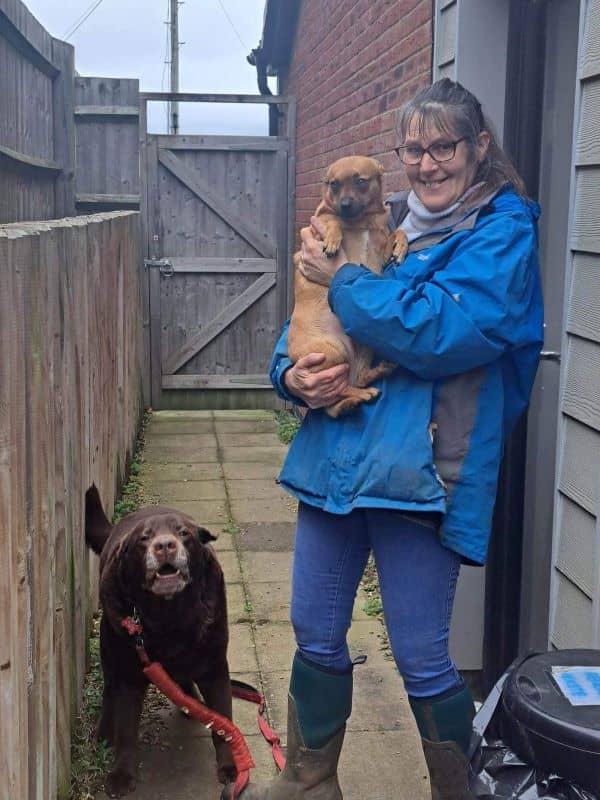 Sarah standing next to a house and carrying a small dog. There is a chocolate lab barking next to her.